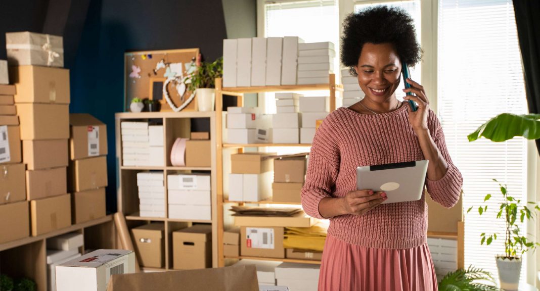 A Black woman and small business owner is in a shop surrounded by packages, looking at her tablet, indicating she might be using the cost of goods sold formula for her accounting.