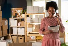 Calcular el costo de los bienes vendidos: guía paso a paso A Black woman and small business owner is in a shop surrounded by packages, looking at her tablet, indicating she might be using the cost of goods sold formula for her accounting.